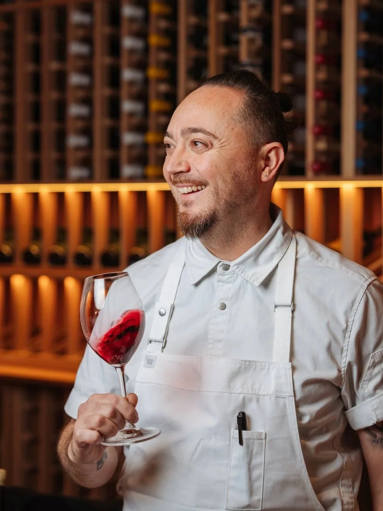 a smiling chef in a white short-sleeve jacket and white canvas apron holding a large crystal wine glass with deep red wine, standing in front of a floor-to-ceiling timber wine rack glowing with warm amber light
