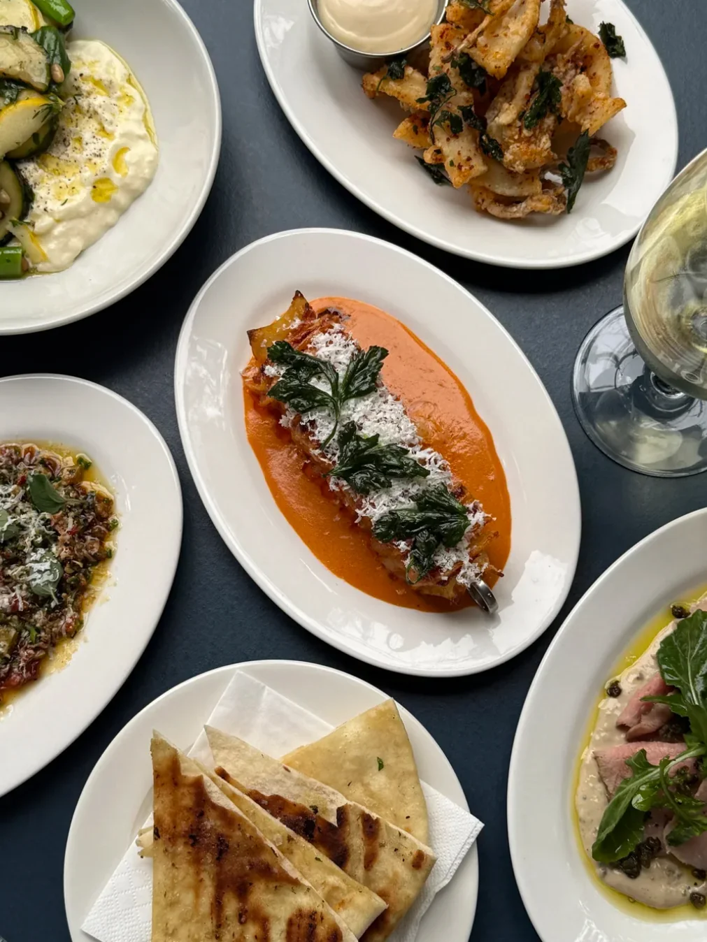 Share plates on a dark table at Fior in Gymea, including grilled flatbread, fried calamari with dipping sauce and a dish in orange sauce with grated cheese.