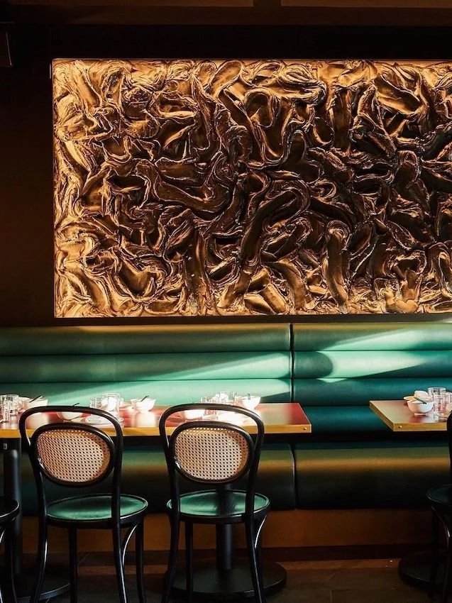 a moody restaurant interior with a deep green leather banquette, black bentwood cane chairs and timber tables set with glassware, beneath a large backlit bronze sculptural relief artwork with swirling textured forms.