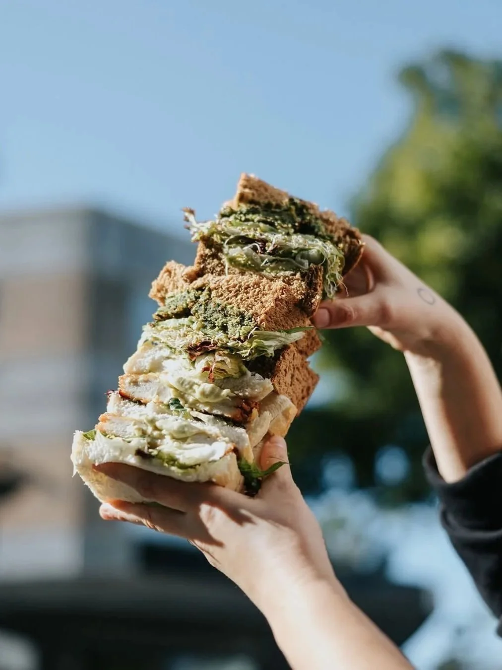 Hands holding a tall stack of sandwiches filled with chicken, greens and pesto against a blurred outdoor background.