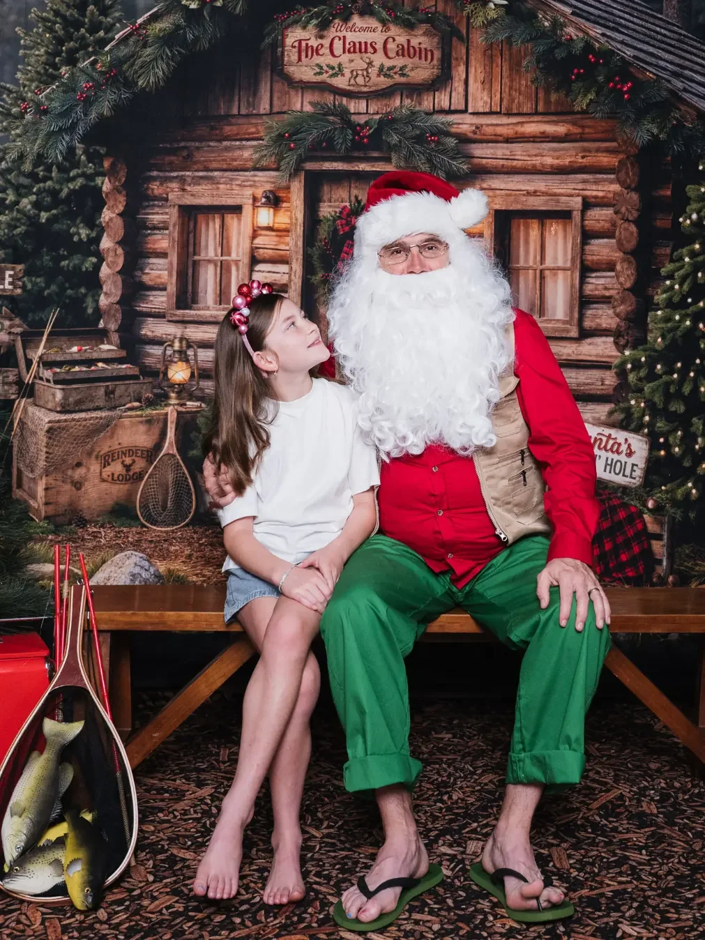 A girl in a sparkly headband looks up at Santa in a log cabin Christmas photo set.