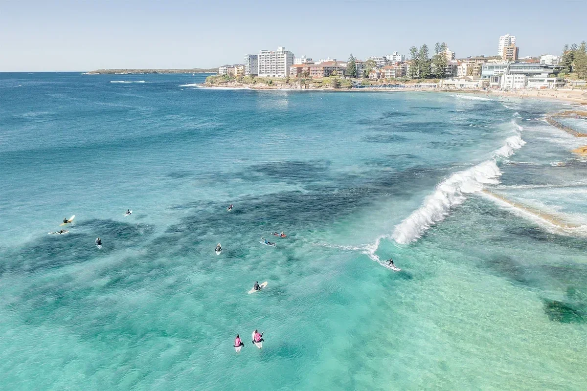 Aerial view of surfers catching waves in crystal-clear turquoise waters at Cronulla Beach, Sydney, with the coastline, sandy shores and coastal buildings in the background on a sunny day.