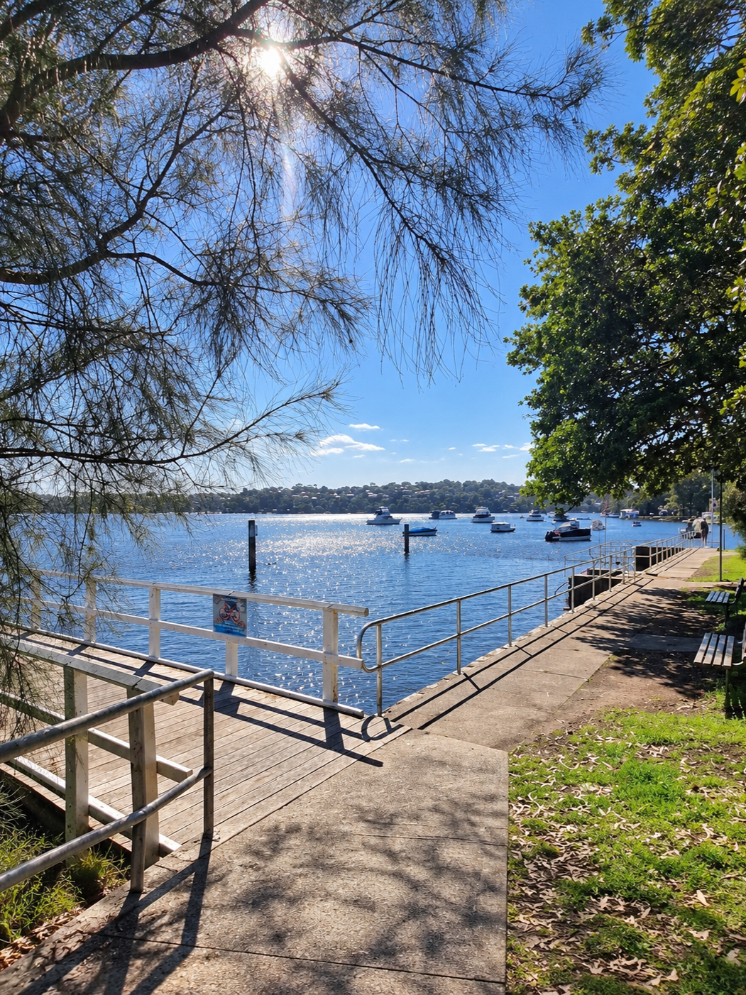 Lilli Pilli Baths in Sydney’s Sutherland Shire, featuring a timber walkway and waterfront swimming enclosure overlooking the calm blue waters of Port Hacking River, with boats anchored in the distance and leafy trees providing shade on a sunny day.