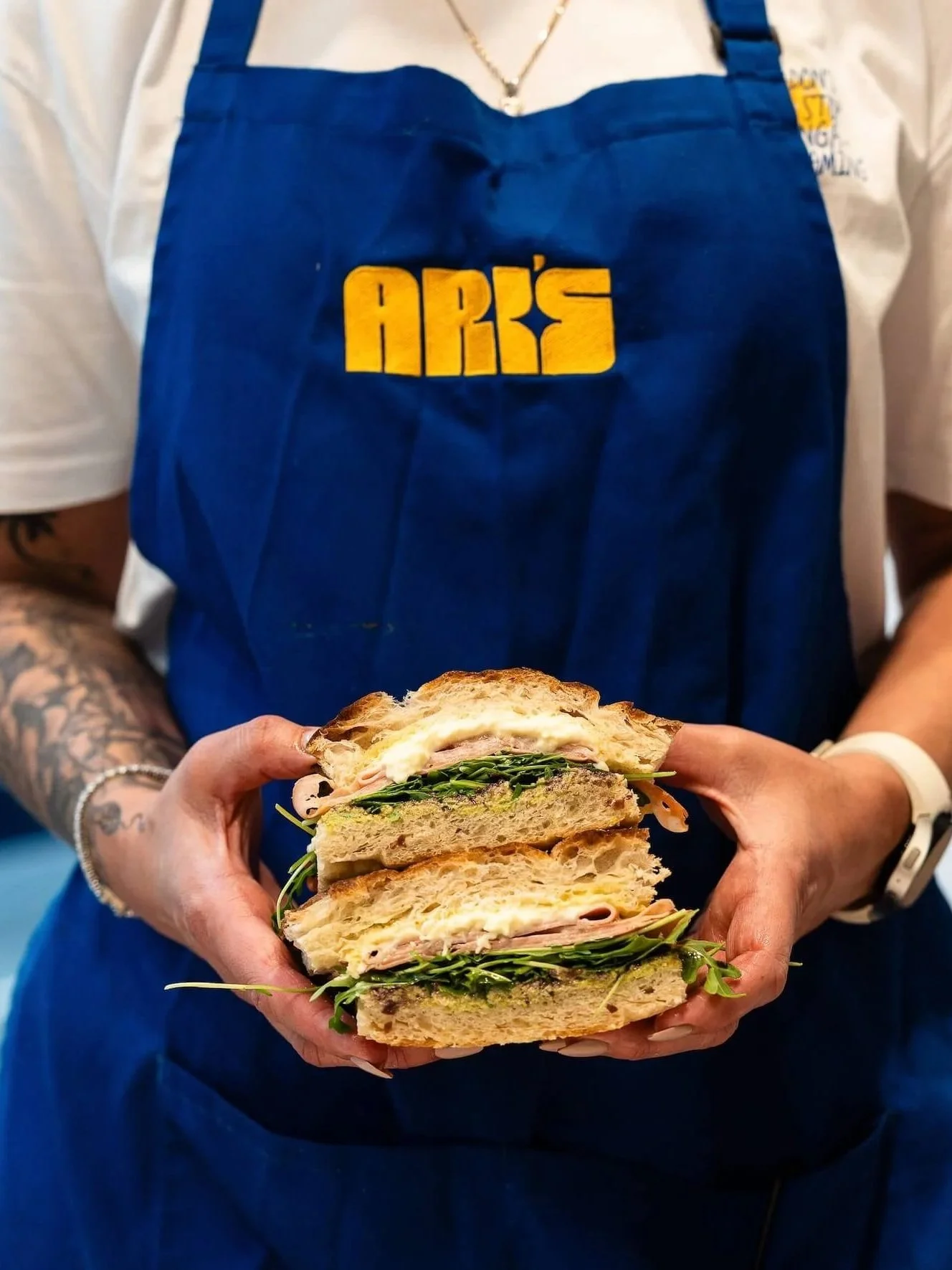 Staff member wearing an Ari’s apron holds a toasted sandwich filled with ham, cheese and rocket.