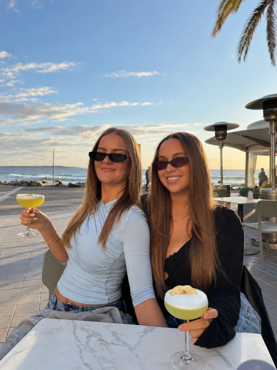 Two women holding cocktails at an oceanfront table at Next Door in Cronulla at sunset.