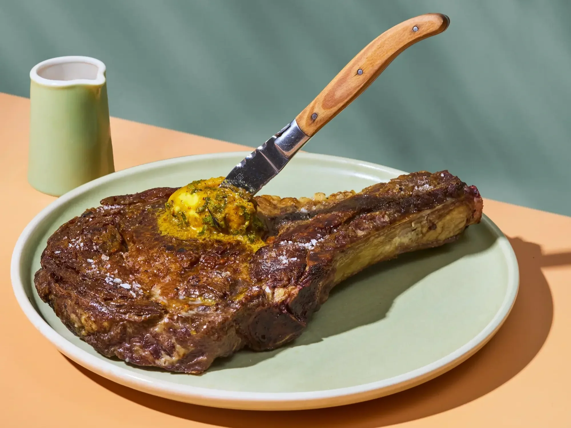 Grilled bone-in rib-eye steak topped with herb butter and sea salt, served on a pale green plate with a steak knife against a green backdrop.