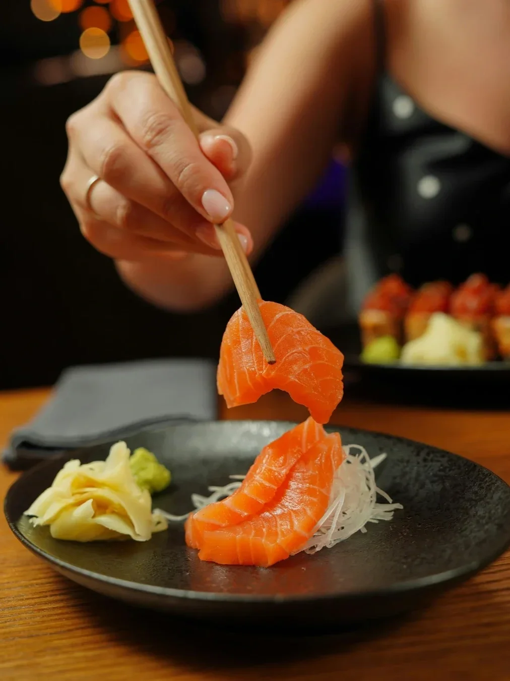Close-up of salmon sashimi being picked up with chopsticks, served with pickled ginger, wasabi and shredded daikon.