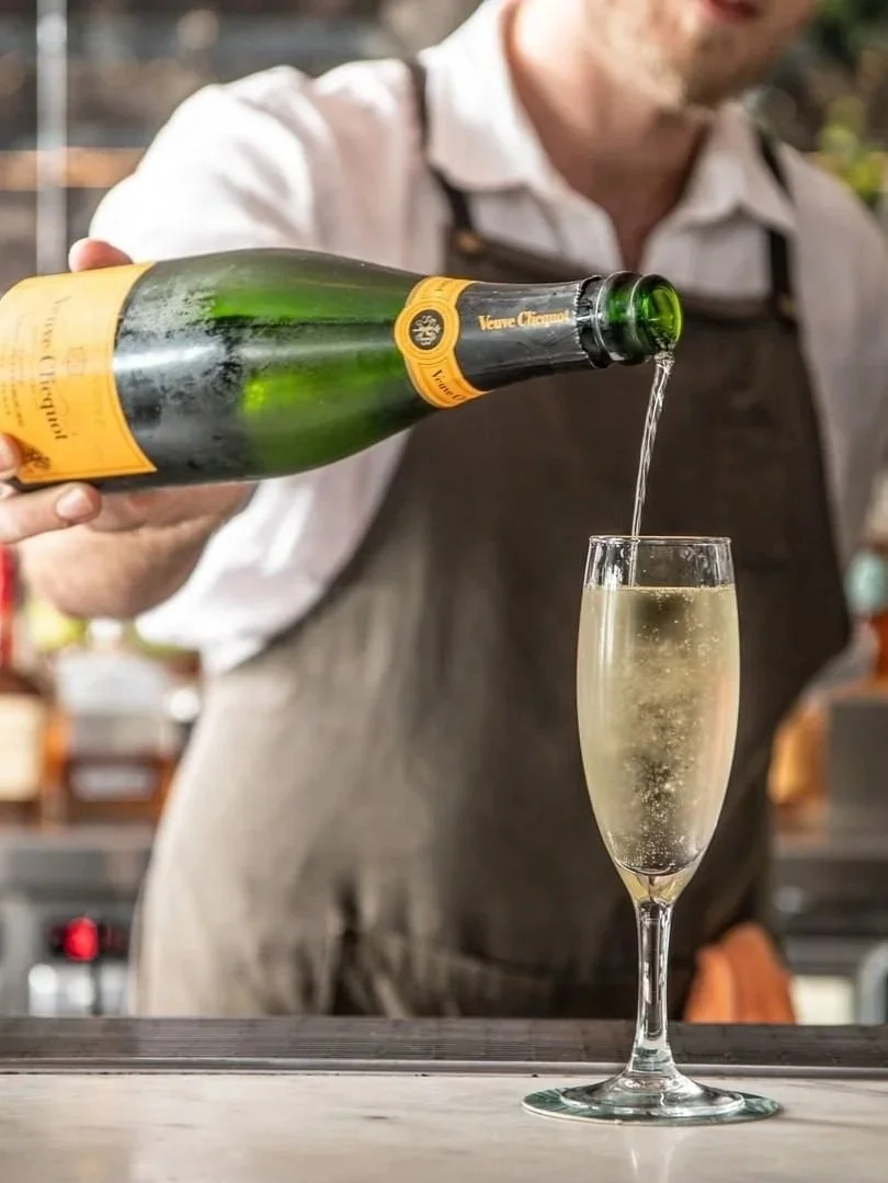 A bartender pouring Veuve Clicquot Champagne into a flute at The Botanical Highfield, featuring a close-up of the champagne bottle, golden bubbles and a stylish bar setting.