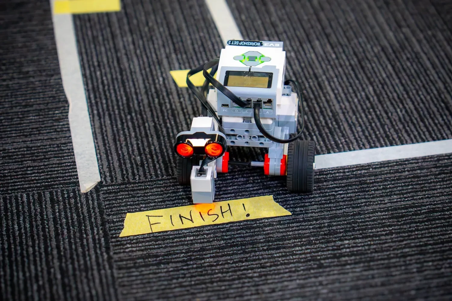 A small programmable robot reaches a handwritten "Finish!" tape marker on a carpeted course at an ANSTO robotics activity.