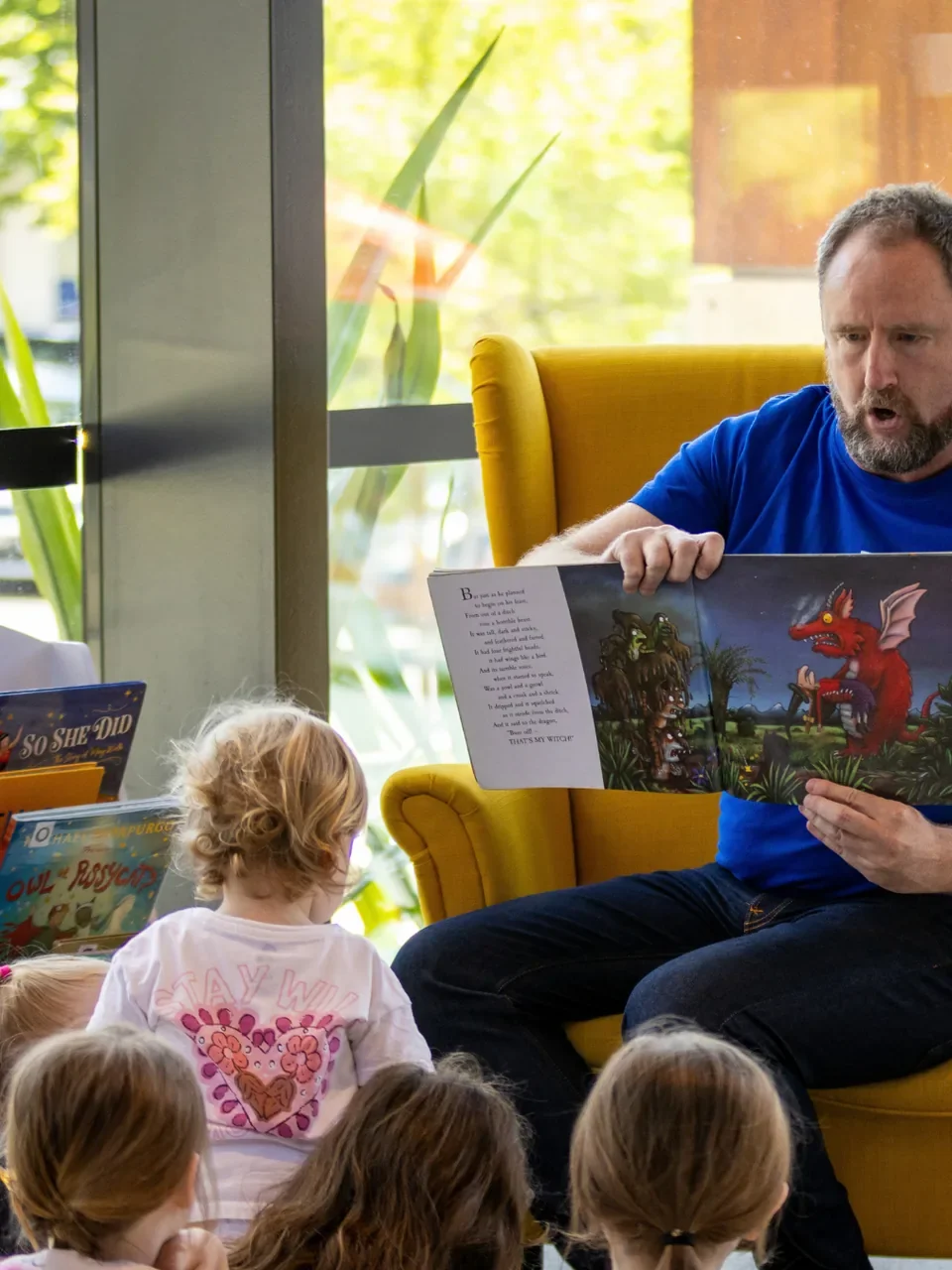 Man reading a picture book aloud from a yellow armchair to young children at Sutherland Pavilion.