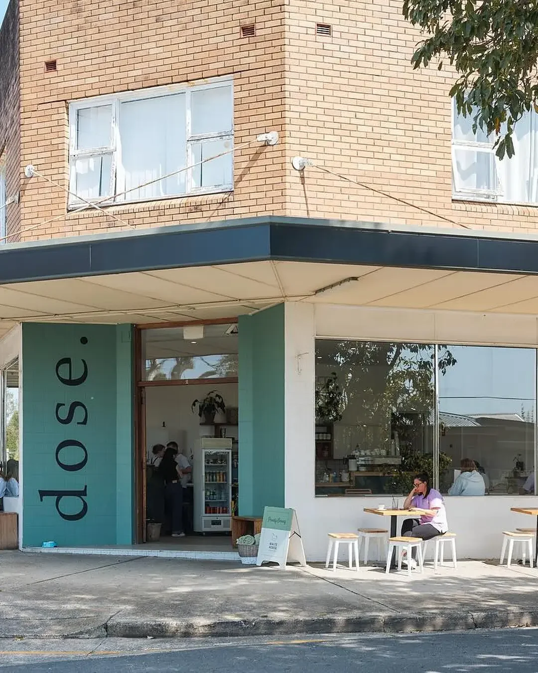 The corner storefront of dose. café in Engadine, with teal tiled cladding, a black awning over tan brick, large glass windows, and white outdoor stools around wooden tables on the footpath.