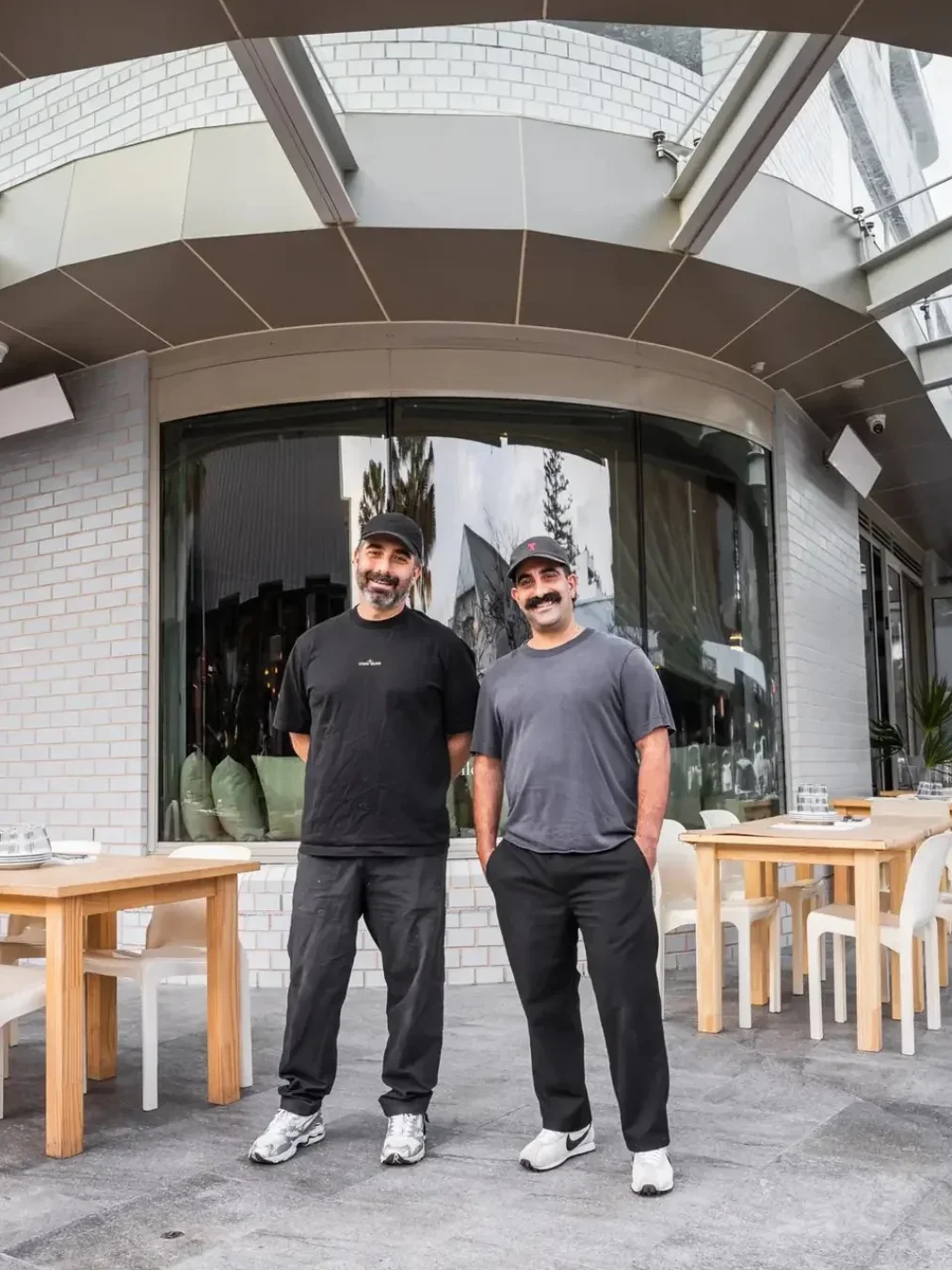 Two men in caps stand smiling in front of the outdoor dining area at Homer in Cronulla.