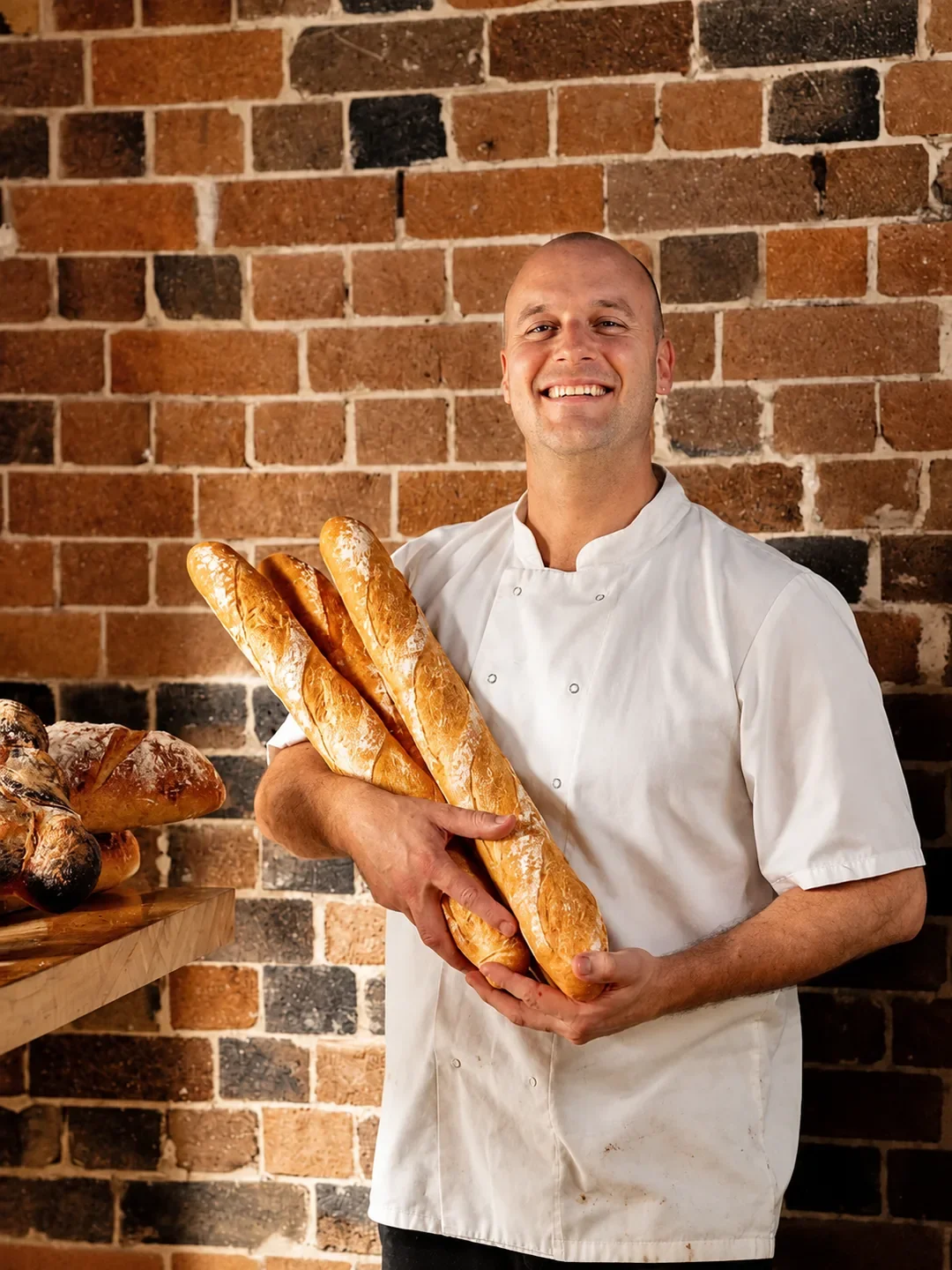 A smiling baker in chef's whites holds an armful of baguettes against a brick wall at Baked by Keiran in Sutherland.