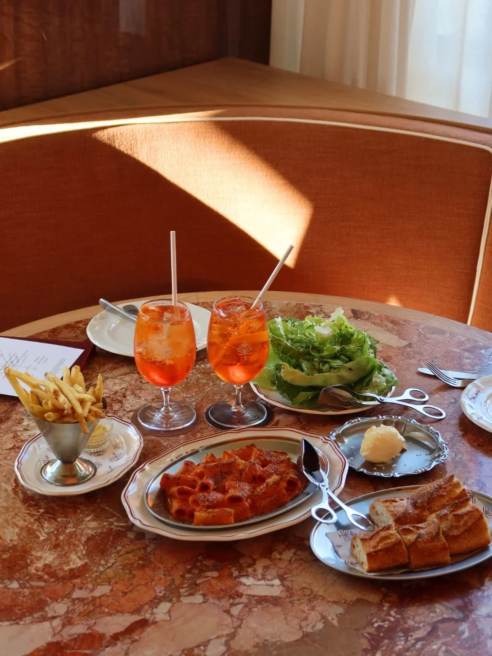 A table spread at Meadow Cronulla with two Aperol spritz cocktails, a baked rigatoni dish, fries in a silver cup, whipped butter, crusty bread and a fresh green salad.