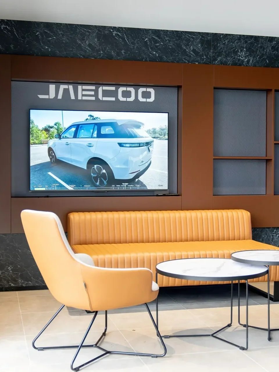 Modern JAECOO dealership lounge with tan leather seating, marble tables, dark stone finishes and a screen displaying a white SUV.