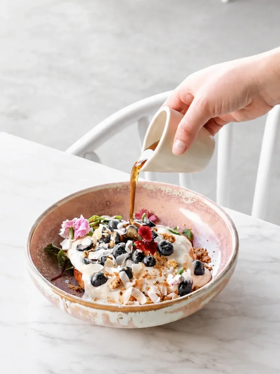 Close-up of syrup being poured over a colourful breakfast dish topped with blueberries, coconut flakes and edible flowers at a café table.