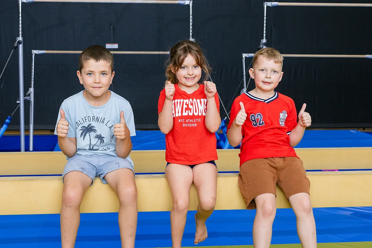Three children sit on a gymnastics beam giving a thumbs up, with uneven bars behind them at PCYC Sutherland.