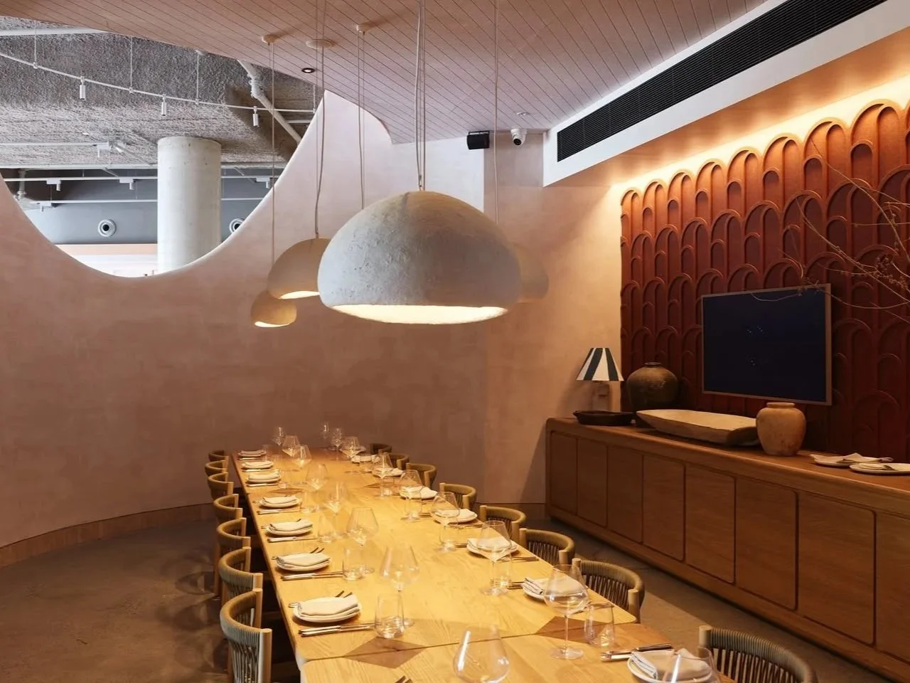 a long timber dining table set with crystal glasses and white napkins in a warm restaurant private dining room with curved plaster walls and oversized concrete pendant lights.