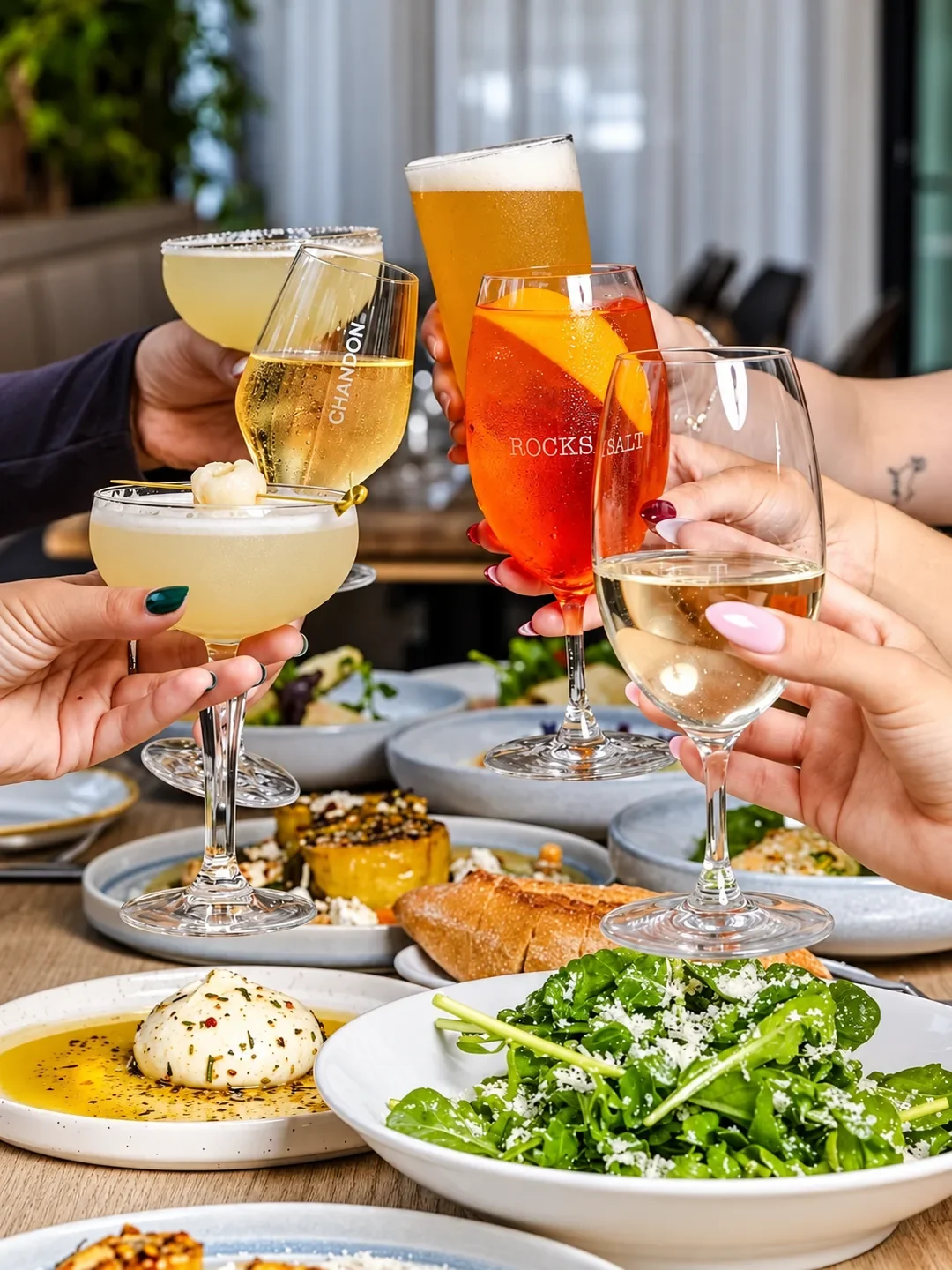 Friends raise cocktails, beer and wine over shared plates, including burrata and a rocket salad, at Rocksalt in Menai.