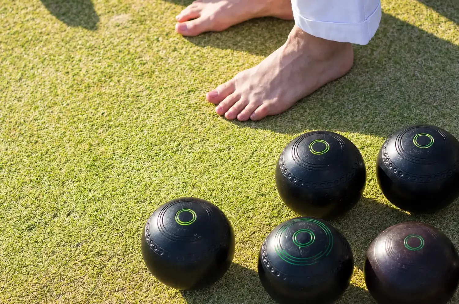 Barefoot bowler standing beside a group of black lawn bowls on the green at Gymea Bowls.