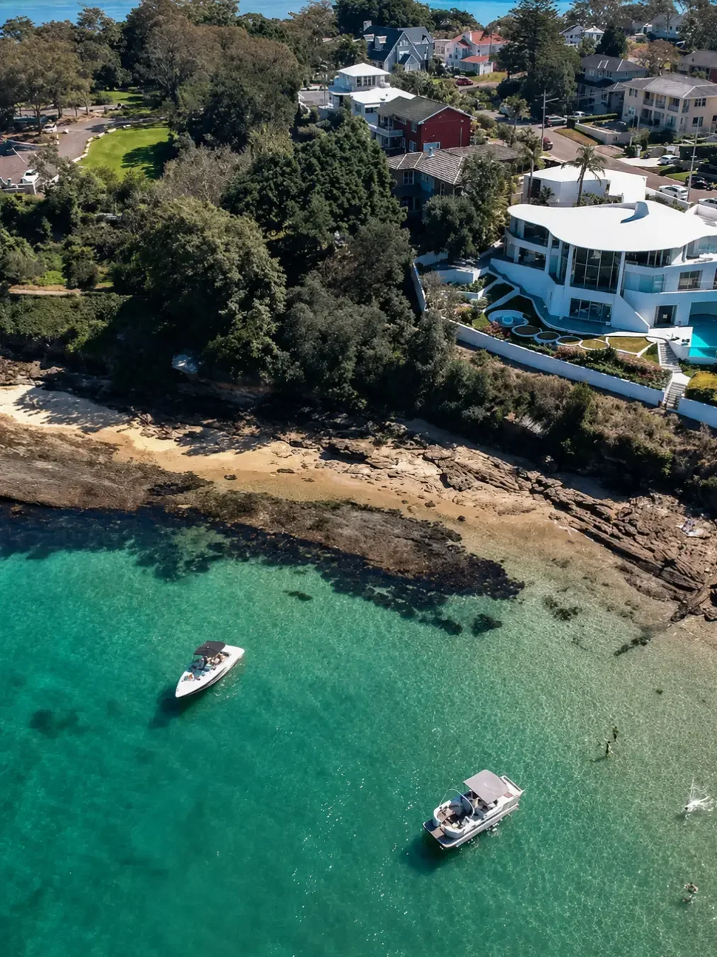 An aerial drone shot of the salmon haul catch site on the Cronulla waterfront, with turquoise water, a rocky sandstone shoreline, and a modern white curved-roof house with a pool overlooking two moored boats.
