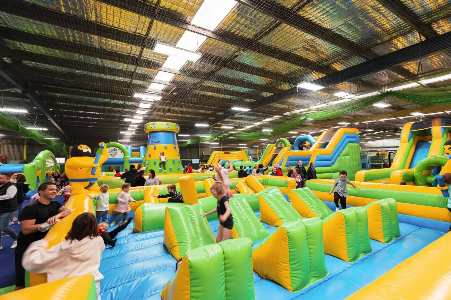 Children play across a large indoor inflatable obstacle course with slides and a climbing tower at Inflatable World Miranda.
