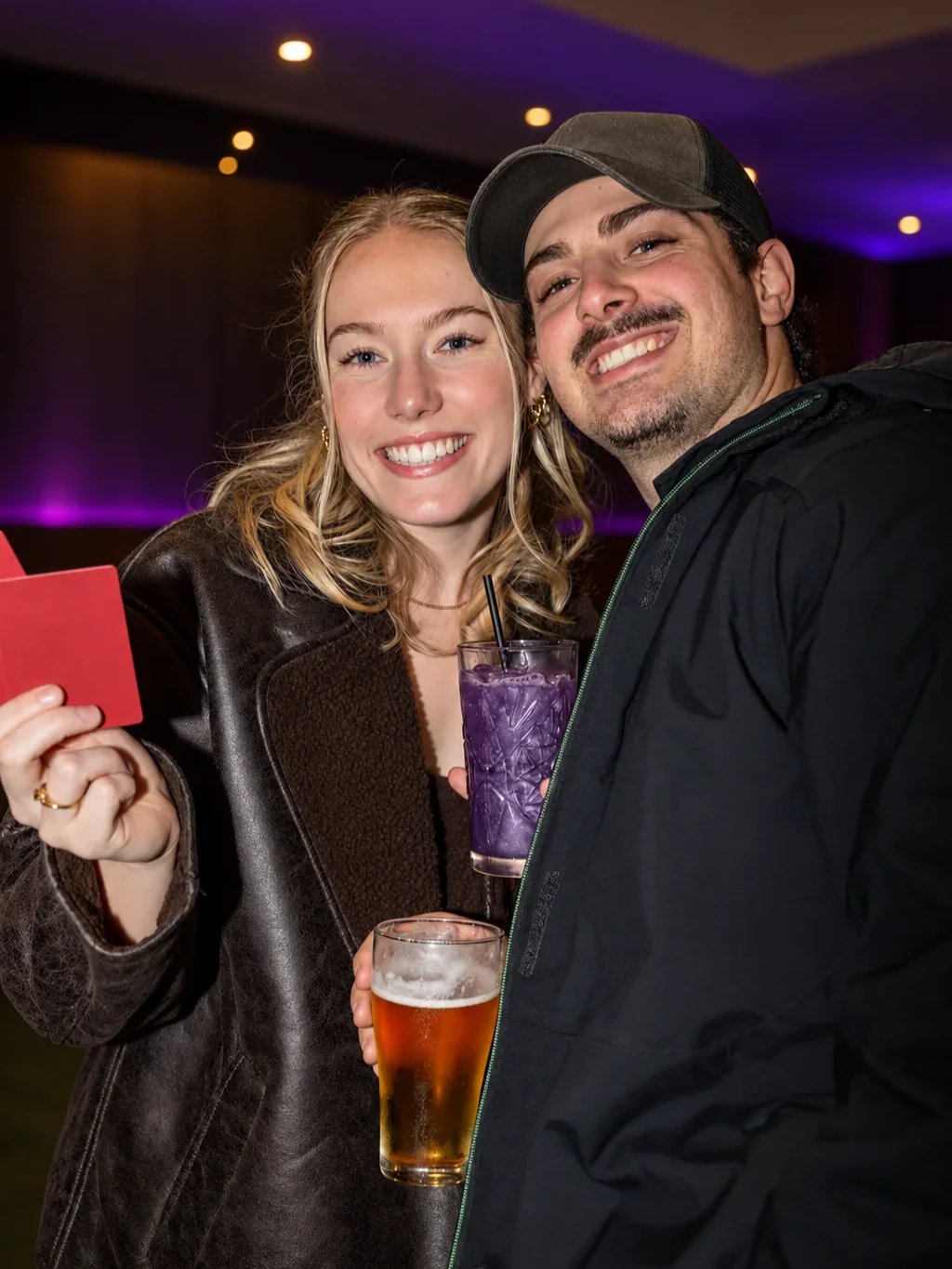 A woman holds up a red card while smiling with a man in a cap, holding a purple cocktail and a beer, at an Infynite Dating Events mixer.