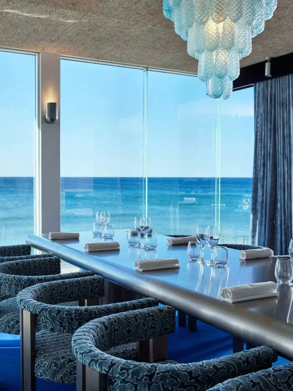 Elegant oceanfront dining room at Summer Salt in Cronulla, featuring a beautifully set table, blue upholstered chairs, sculptural pendant lighting and floor-to-ceiling windows overlooking the ocean.