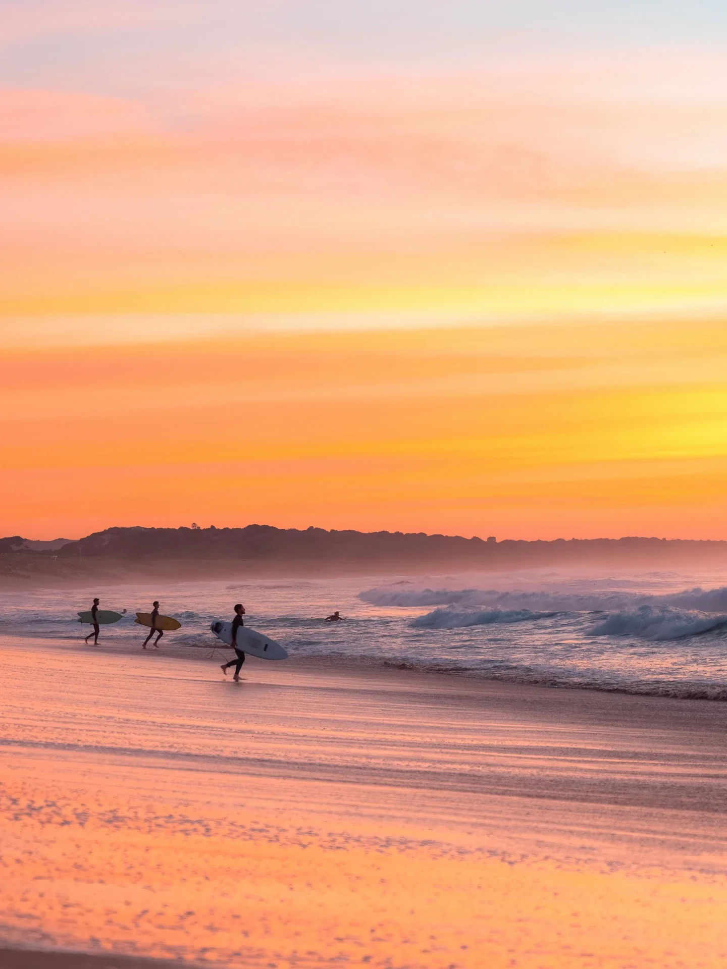 Wanda Beach in Cronulla, Sydney, at sunrise, featuring surfers carrying their boards along the golden sandy shoreline, gentle ocean waves and a vibrant pink, orange and yellow sky reflecting across the wet sand.