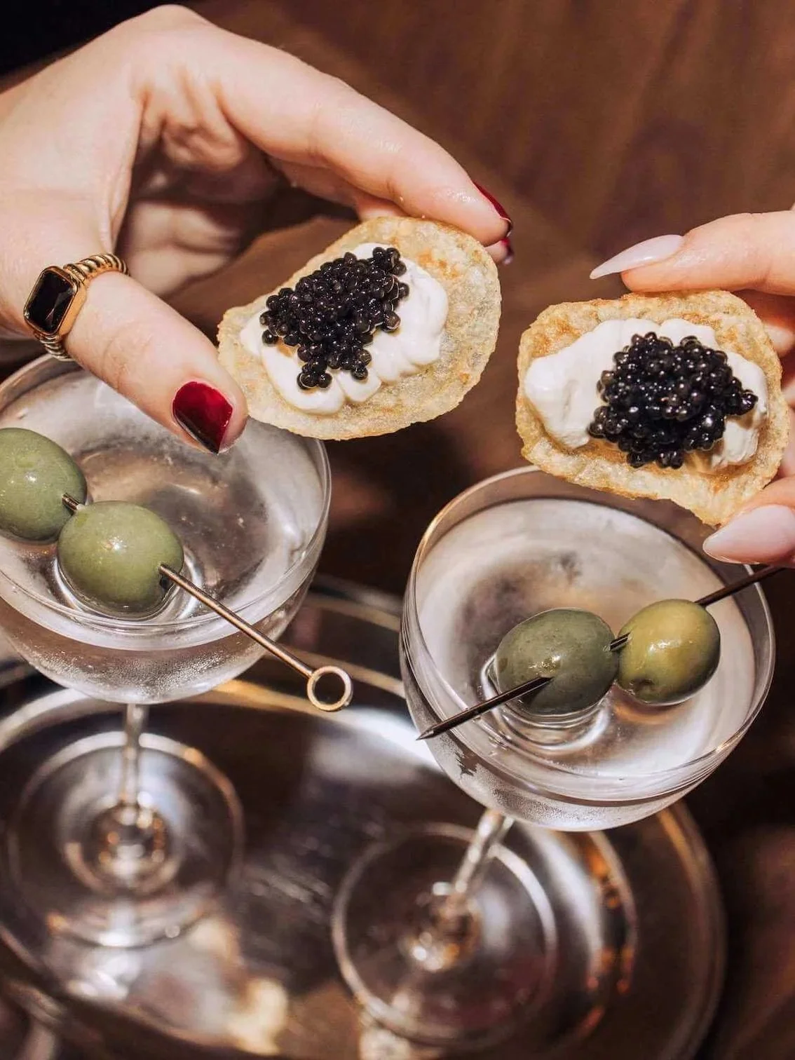 two hands holding blini topped with crème fraîche and black caviar over chilled dirty martinis garnished with green olives on gold cocktail picks, served on a silver tray against a warm dark timber background.