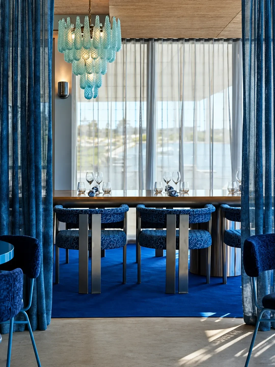 A private dining area at SummerSalt with blue upholstered chairs, a glass chandelier and sheer curtains over water views.