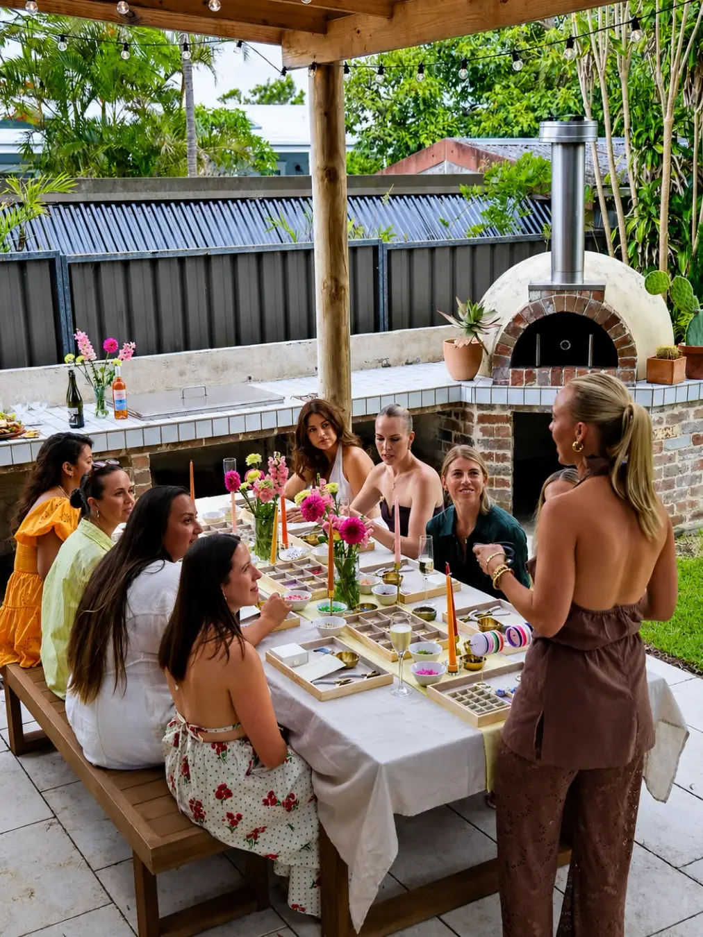 A group of women gathered around an outdoor table at a jewellery-making workshop, with bead trays, candles and pink dahlias, a wood-fired pizza oven and string lights in the background.