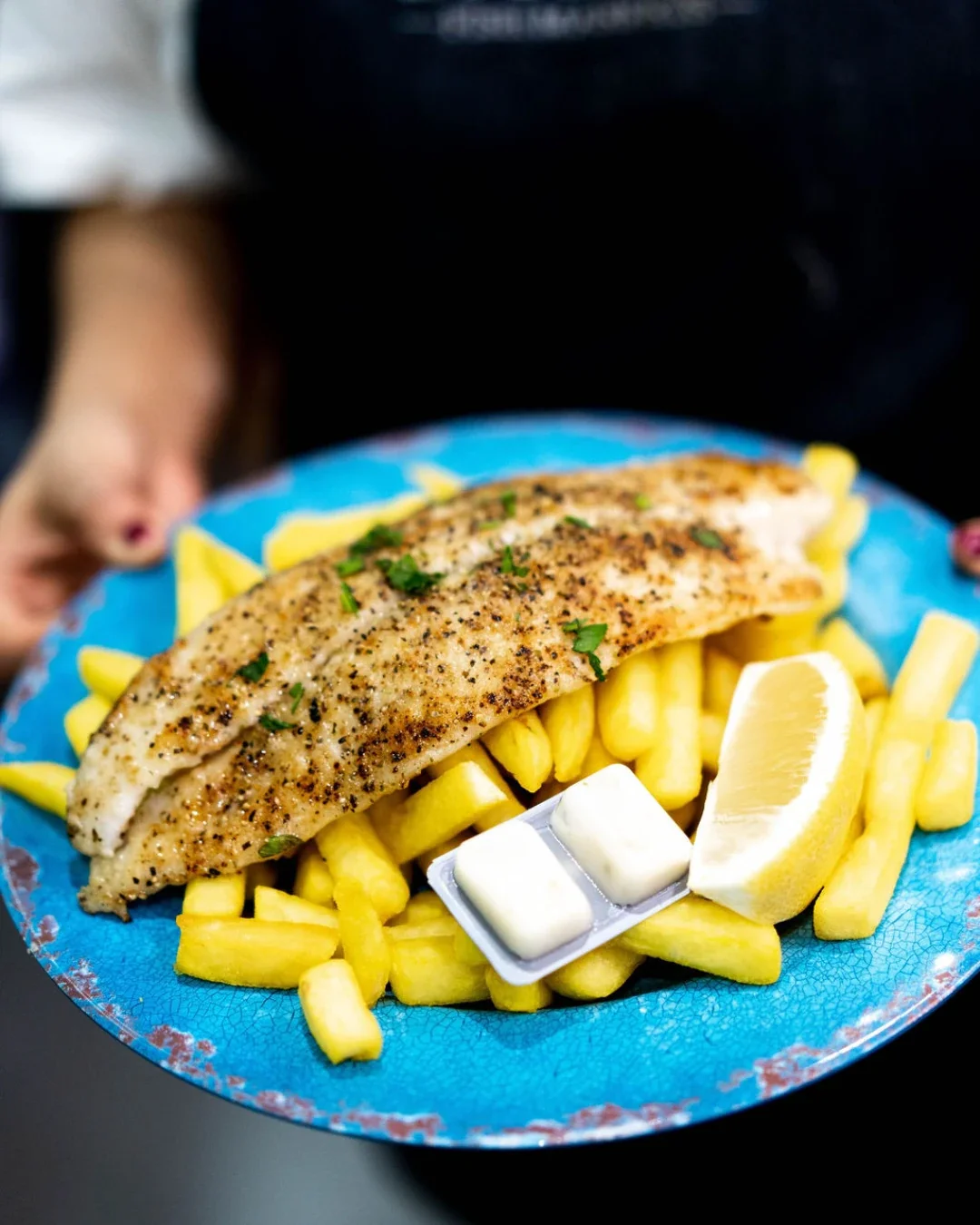 A grilled fish fillet dusted with cracked pepper and fresh parsley, served on a rustic blue ceramic plate with golden chips, a lemon wedge and two portions of butter, held out by a person in a black apron.