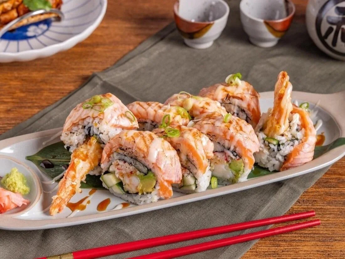 a white oval platter of prawn and salmon sushi rolls drizzled with teriyaki sauce, served with wasabi, pickled ginger and red chopsticks on a grey linen napkin.