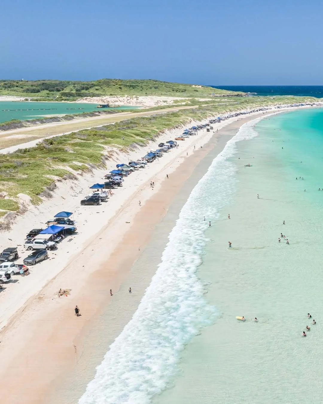 Aerial view of Boat Harbour Beach in Kurnell, Sydney, featuring a long stretch of white sand lined with four-wheel drives and beach shelters, swimmers enjoying clear turquoise waters and gentle waves along the coastline on a sunny day.