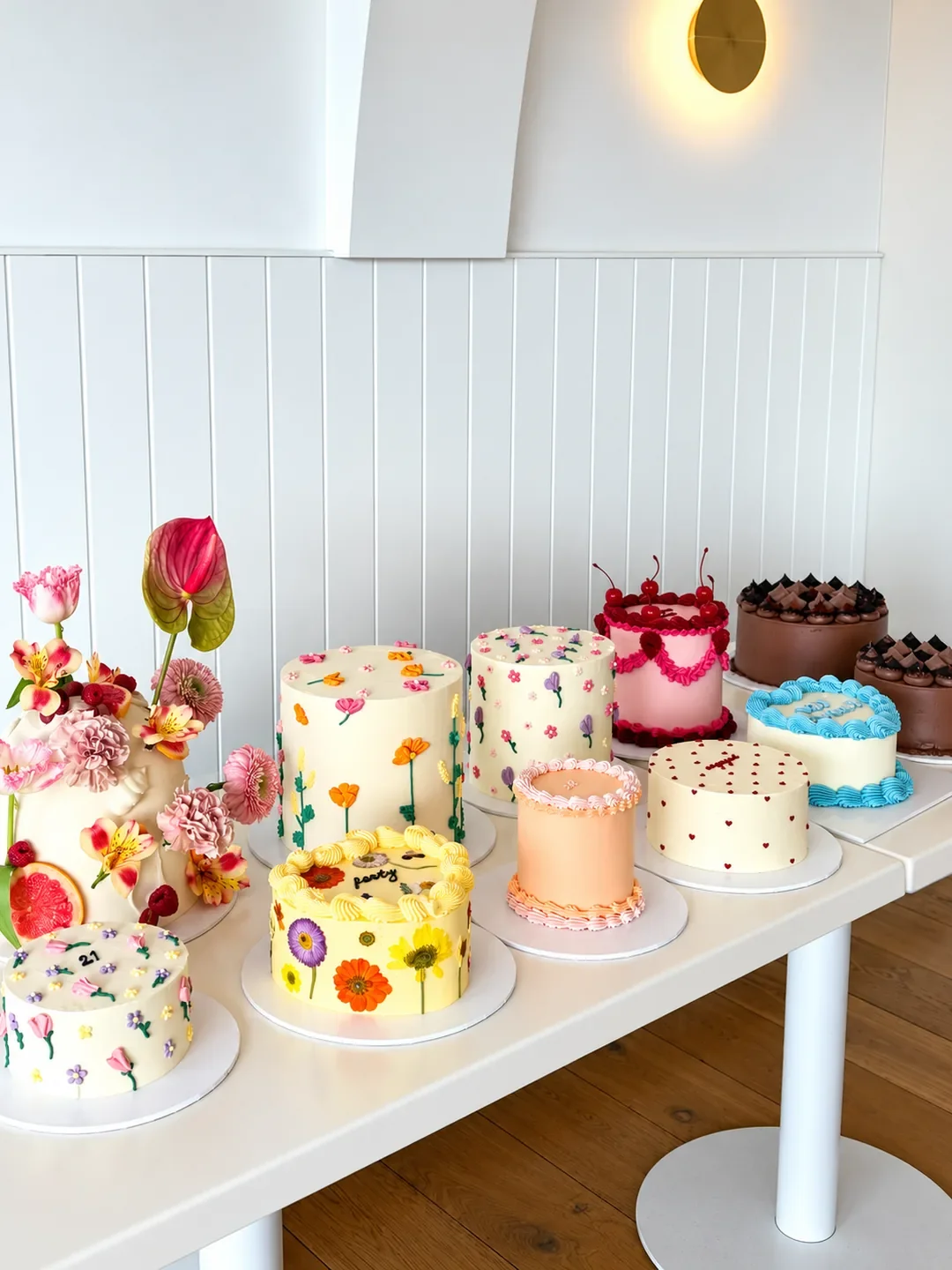A table of decorated celebration cakes at Baked Collective in Caringbah, including floral buttercream designs, cherry-topped pink and chocolate cakes.