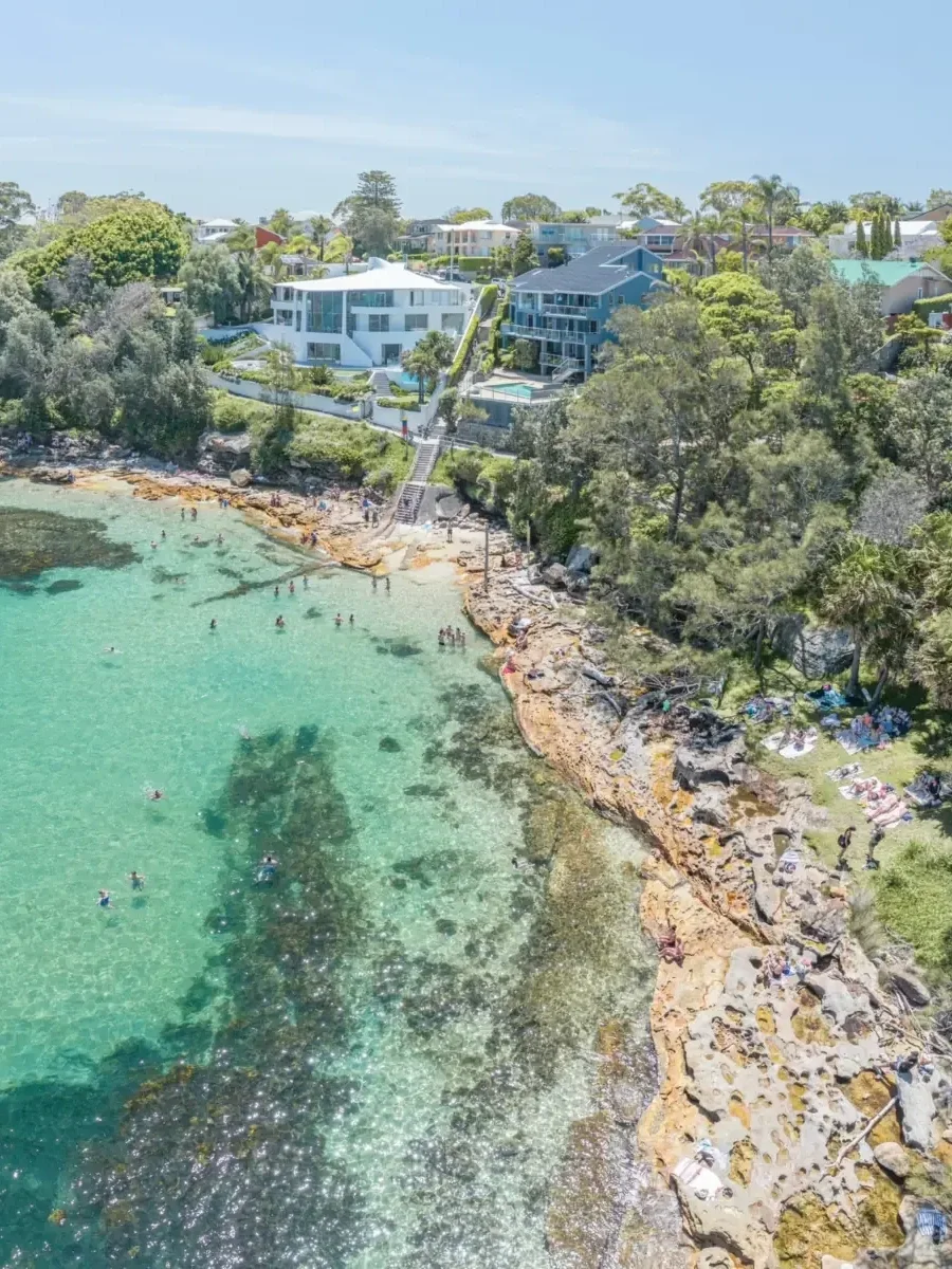 Aerial view of Salmon Haul in Cronulla, Sydney, featuring crystal-clear turquoise waters, swimmers enjoying the sheltered bay and sunbathers relaxing along the rocky shoreline, with lush greenery and coastal homes overlooking the water.