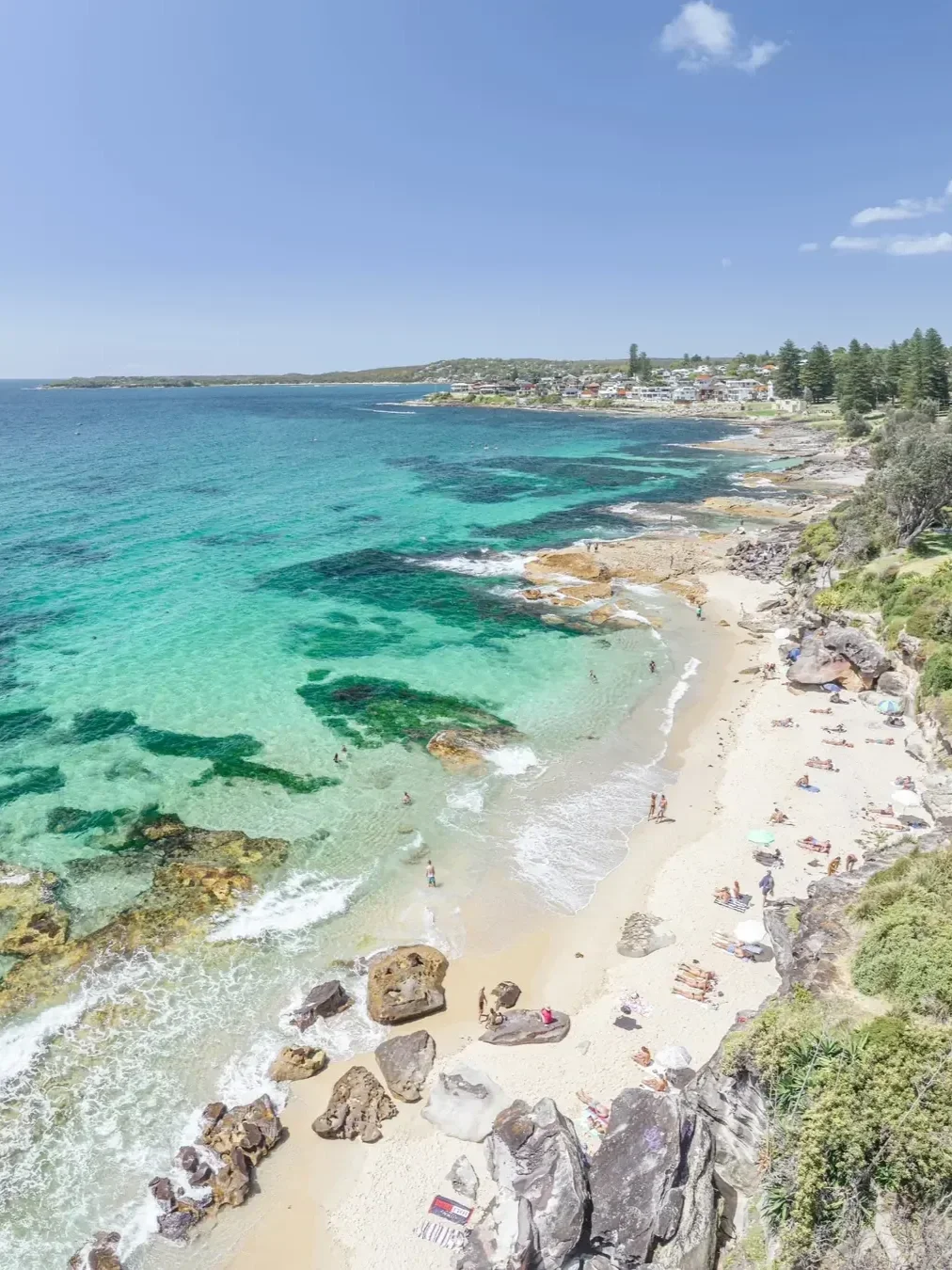 Aerial view of Blackwoods Beach in Cronulla, featuring turquoise waters, golden sand, sandstone rocks and beachgoers enjoying a sunny day.