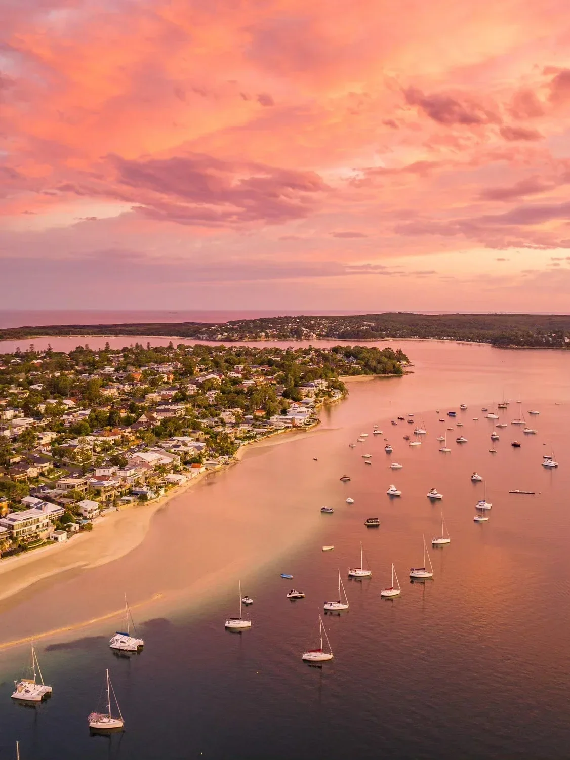 Aerial view of Gunnamatta Bay in Cronulla, Sydney, at sunset, featuring calm waters dotted with anchored boats, waterfront homes along the shoreline and a vibrant pink and orange sky reflecting across the bay.