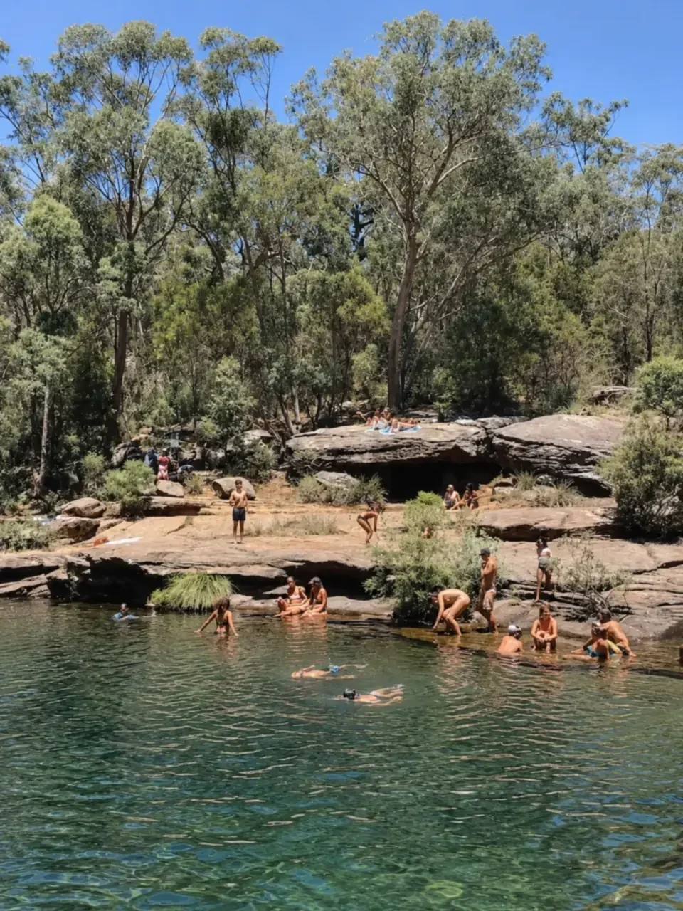 Karloo Pools in the Royal National Park, Sydney, featuring a natural bush swimming hole surrounded by sandstone rocks and lush eucalyptus trees, with visitors swimming and relaxing by the water on a sunny day.