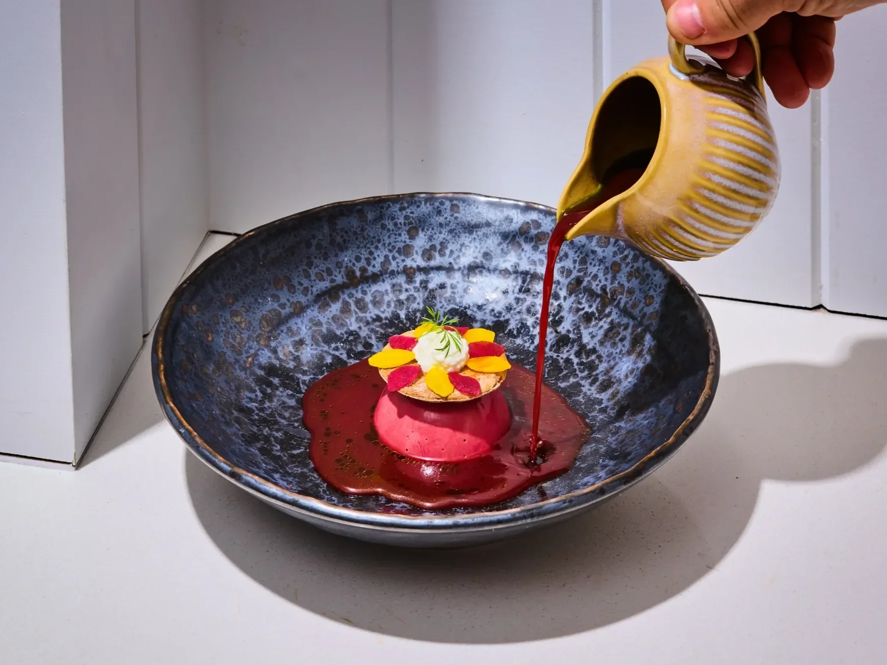 Beetroot dish topped with goat’s curd, edible flowers and herbs, served in a blue ceramic bowl as a red sauce is poured over the plate.