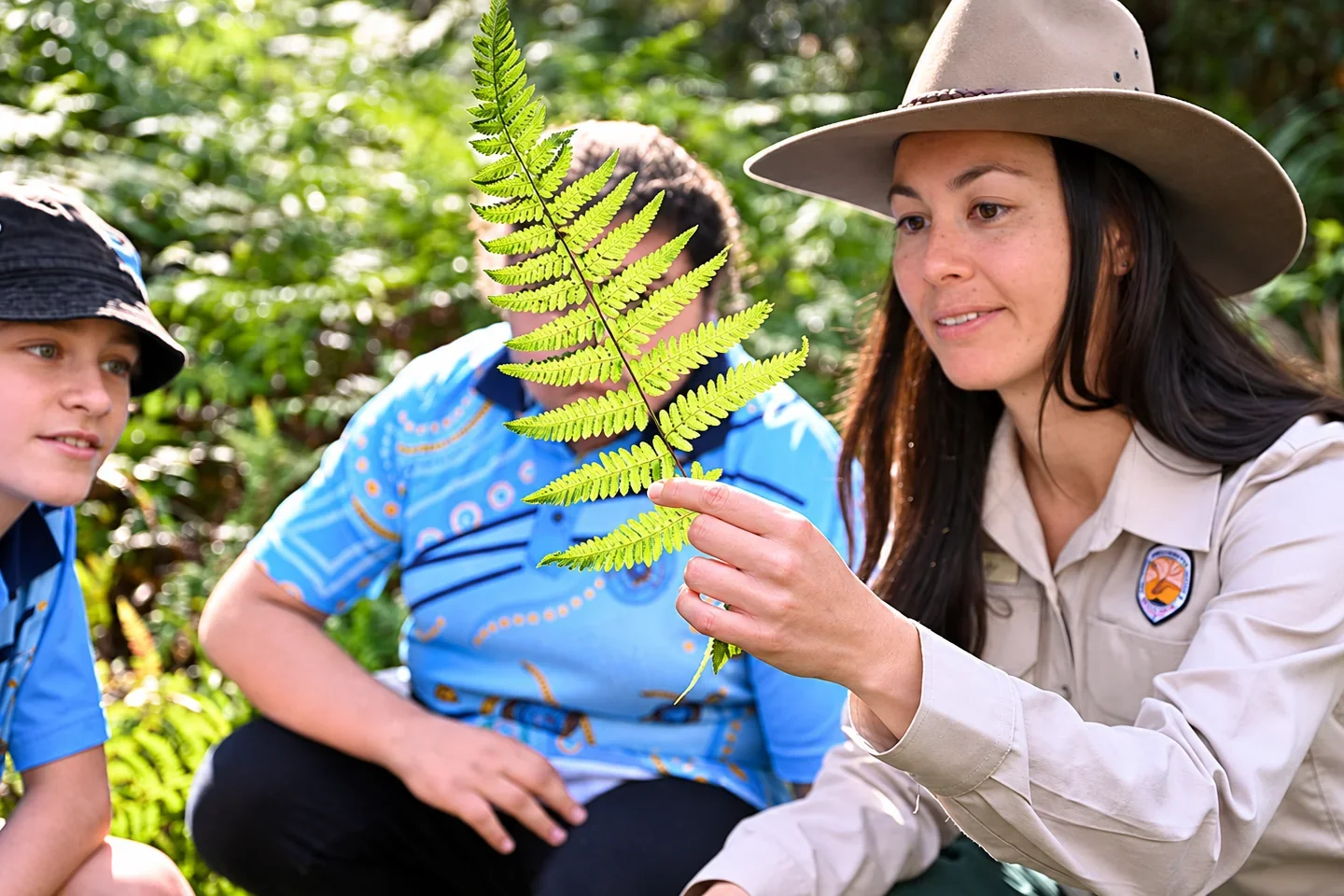 A national parks ranger holds up a fern frond for two children to look at in leafy bushland.