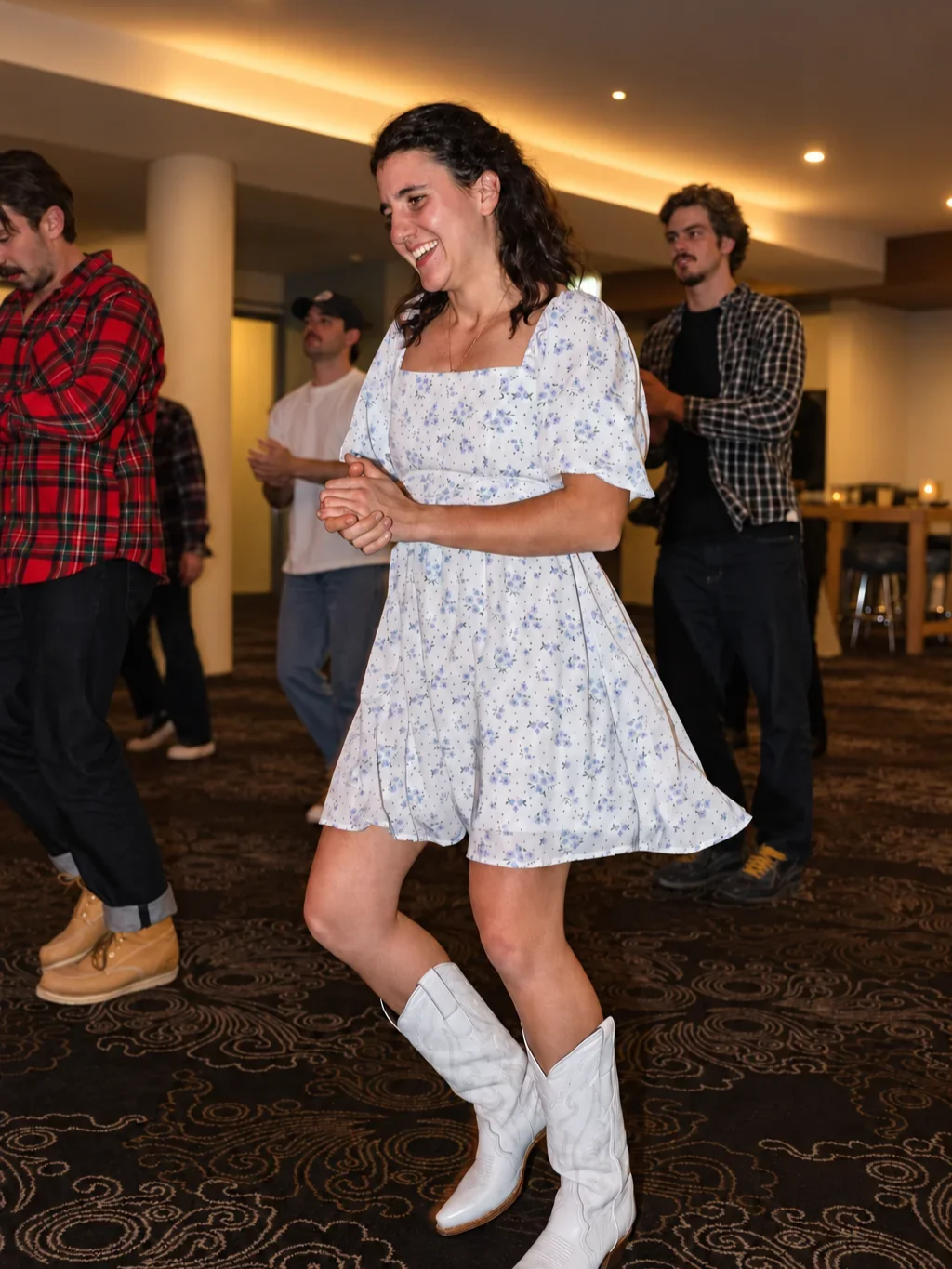 A woman in a floral dress and white cowboy boots laughs while dancing at an Infynite Dating Events cowboy-themed party.