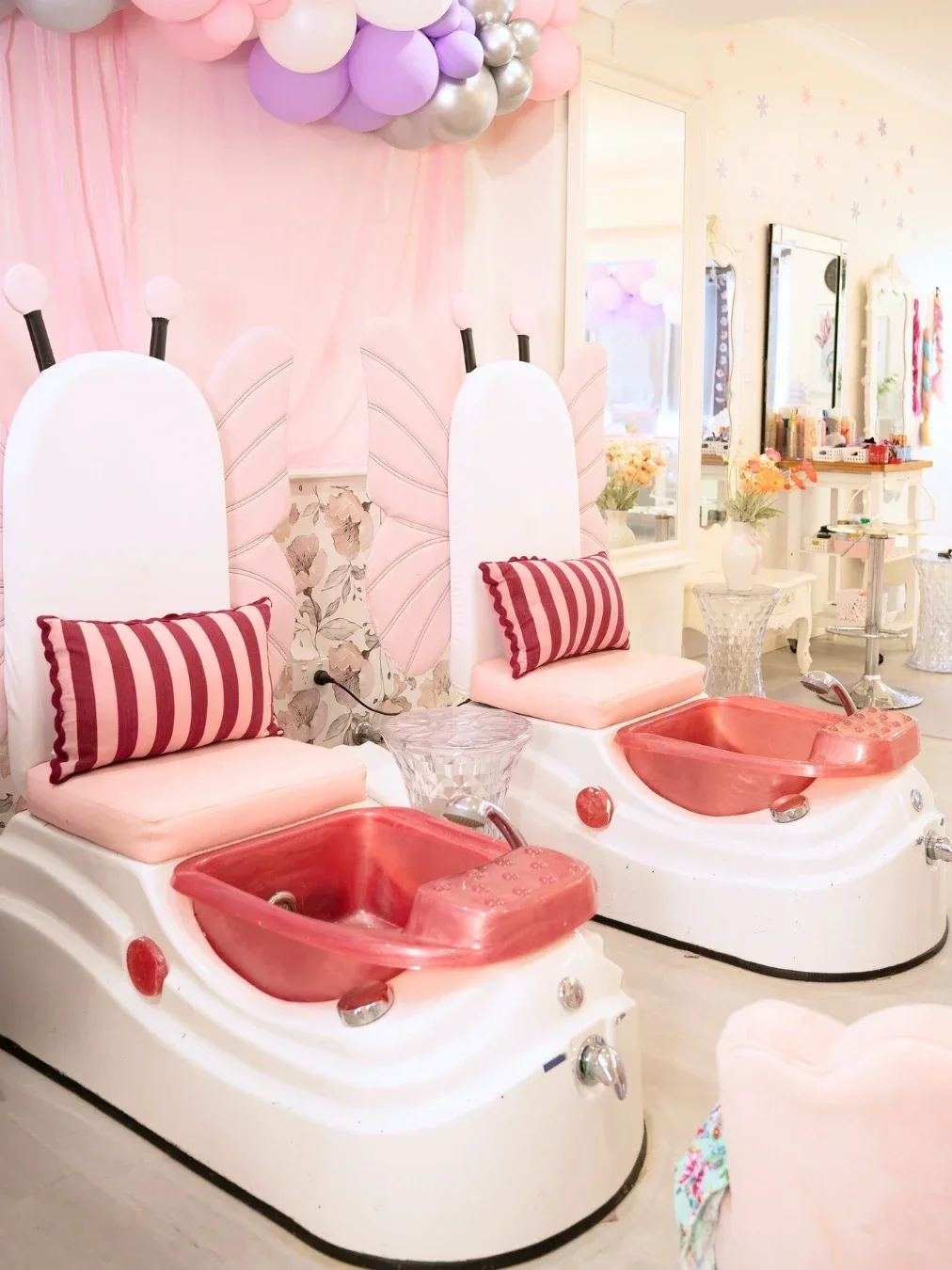 Pink children’s pamper parlour interior with two spa pedicure chairs, striped cushions, pastel balloons and beauty stations.