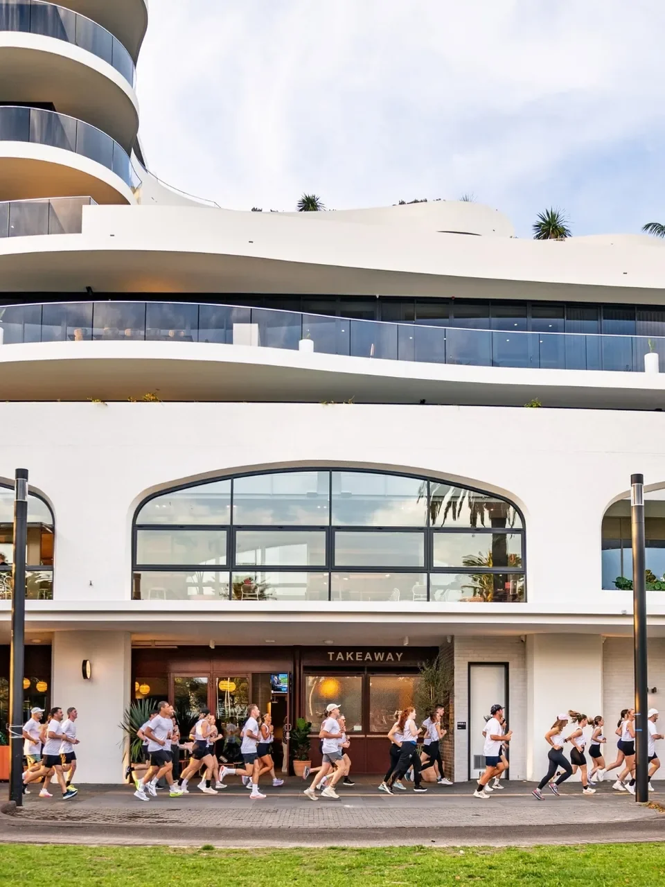 Group of runners in white tops passing the curved white Parc building in Cronulla.