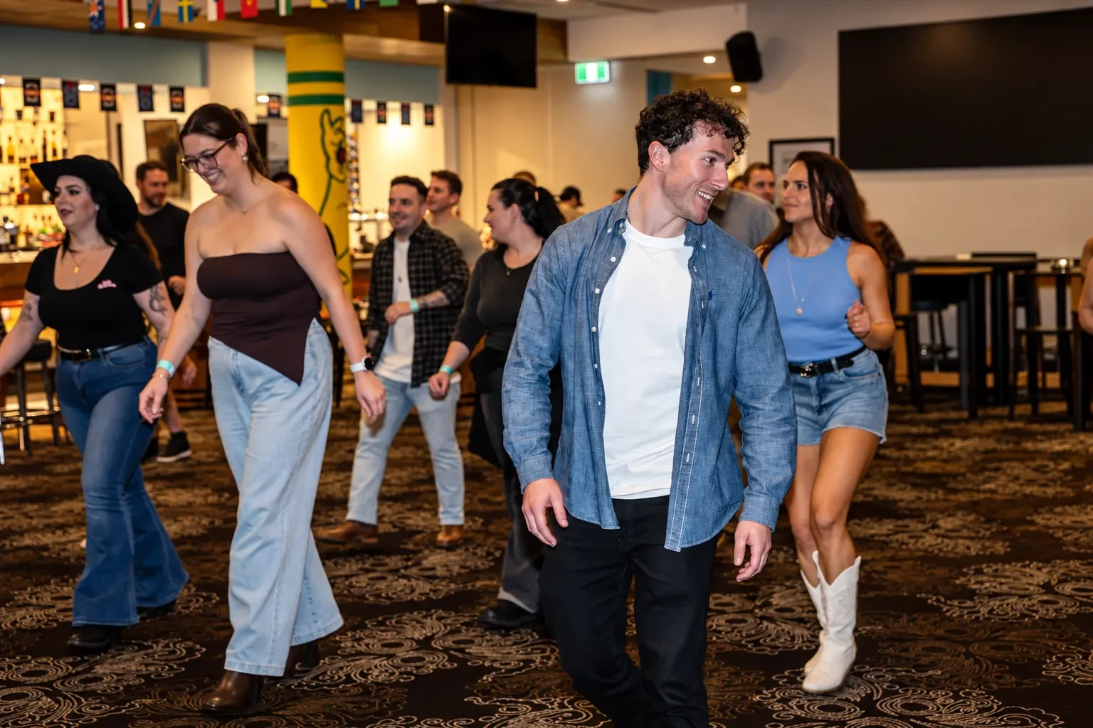 A man in a denim shirt leads a line of dancers on the dance floor at an Infynite Dating Events cowboy-themed party.