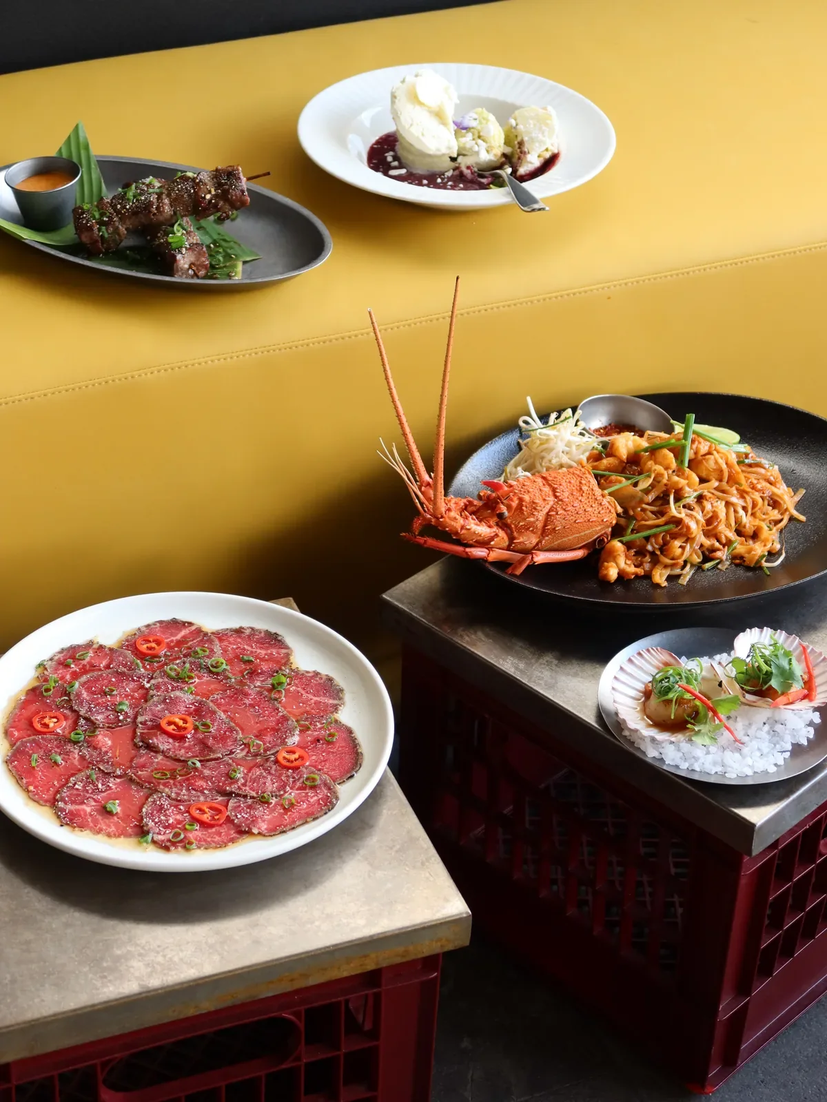 A vibrant Thai-inspired feast at Alphabet St. in Cronulla, featuring lobster noodles, thinly sliced beef, seafood dishes, grilled skewers and a coconut dessert, arranged across bright yellow tables and industrial-style crates.