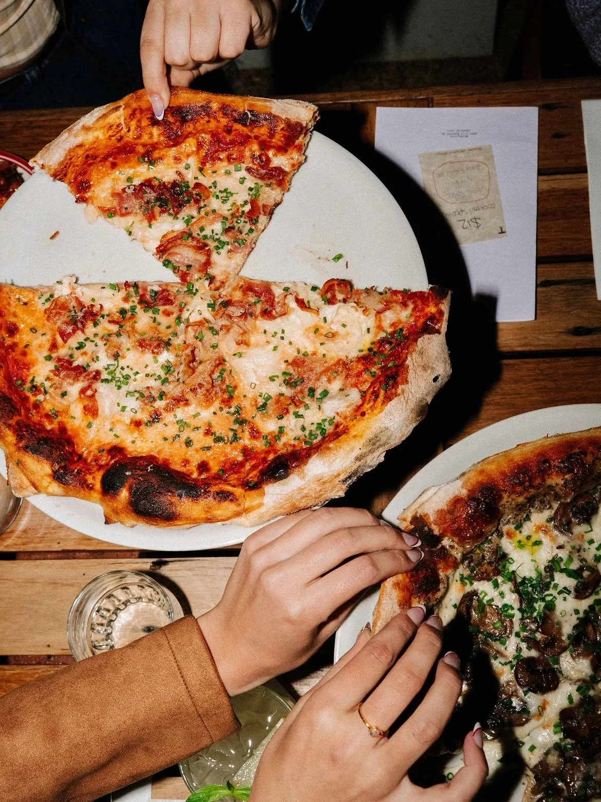 Overhead view of diners reaching for slices of freshly baked pizza topped with cheese, bacon, mushrooms and herbs on a rustic wooden table.