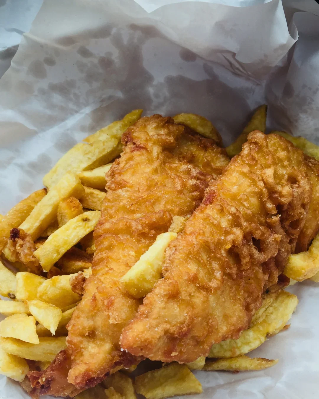 Two golden beer-battered fish fillets with a crisp, bubbled crust, dusted with salt, served on a generous bed of crinkle-cut chips wrapped in white takeaway paper.