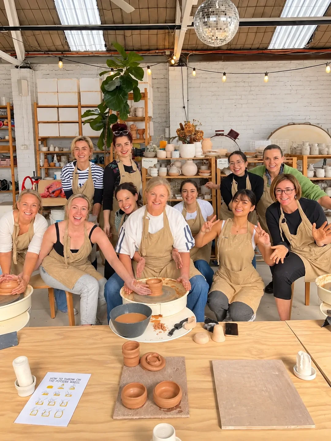A group in aprons smiles around a pottery wheel during a class at Part Time Ceramics in the Sutherland Shire.